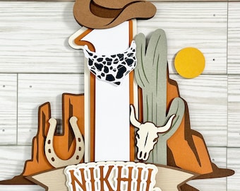 May include: A brown cowboy hat sits atop a white number one cake topper. The number one is decorated with a black and white bandana. The cake topper also features a brown horseshoe, a white skull with horns, and a green cactus. The cake topper is decorated with the name "NIKHIL" in brown letters.