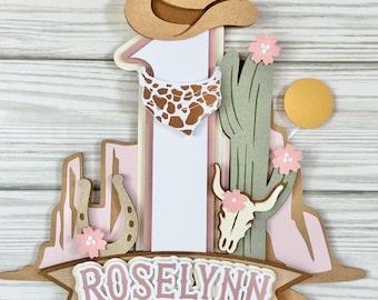 Puede incluir: Adorno de pastel de papel en capas con temática del oeste. Presenta un sombrero de vaquero, pañuelo, cactus, herradura y una calavera de toro. El nombre "ROSELYNN" se muestra en una pancarta. La paleta de colores incluye tonos de marrón, rosa y verde.