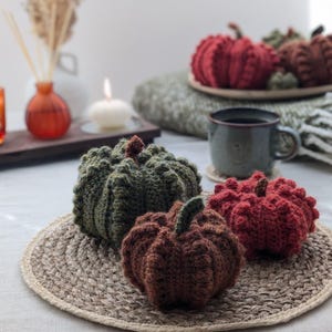 May include: Three crocheted pumpkins in dark green, brown, and deep red colours. Each pumpkin features a textured, ribbed surface with a small stem on top. The pumpkins are arranged on a round, woven jute placemat, showcasing detailed yarn work.