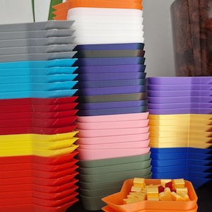 May include: A collection of brightly coloured, geometric trays in shades of red, yellow, blue, and purple. The trays are stacked, highlighting their angular design. Some orange trays hold small, yellow and red objects.