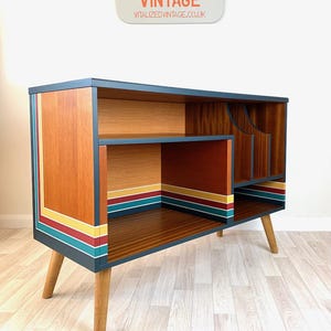 May include: A vintage wooden cabinet with a dark blue frame and legs. The cabinet has a warm wood grain finish with colourful stripes of yellow, red, and teal. The sign above reads "VITALIZED VINTAGE" and "VITALIZEDVINTAGE.CO.UK".