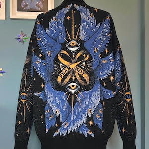 Peut inclure: Veste de bombardier noire avec un motif bleu et or saisissant dans le dos. Le motif présente des ailes stylisées, des yeux et les mots "FEAR NOT". La veste a un col, des poignets et un ourlet côtelés.