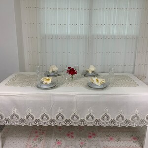 Puede incluir: Un mantel blanco con un delicado borde de encaje y un centro de mesa floral. El mantel está puesto para una cena formal con cuatro cubiertos, cada uno con una servilleta blanca doblada en forma de abanico y un vaso transparente.