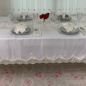 Puede incluir: Mantel blanco con ribete de encaje, preparado para una cena formal con platos blancos, cubiertos y vasos. Un pequeño jarrón con flores rojas se encuentra en el centro de la mesa.