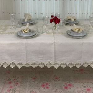 Puede incluir: Un mantel rectangular blanco con un borde de encaje decorativo, preparado para una cena formal. La mesa está puesta con platos, servilletas, vasos y un pequeño jarrón con flores rojas.