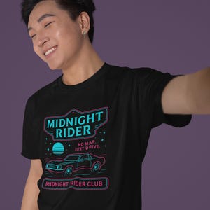 May include: Black t-shirt with a neon blue and pink graphic design of a car with the text "Midnight Rider" and "No map, just drive."  The graphic also includes stars and a crescent moon. The text "Midnight Rider Club" is at the bottom of the graphic.