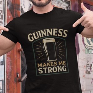May include: Black t-shirt with the text "GUINNESS MAKES ME STRONG" in a cream-colored font. The design includes an illustration of a pint of Guinness. The person in the image is pointing at the shirt.