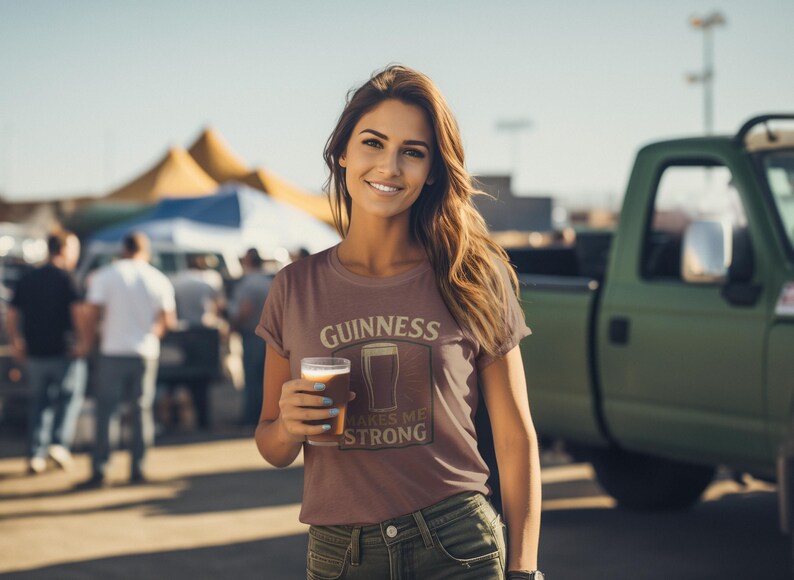 May include: A woman wearing a brown t-shirt with the Guinness logo and holding a pint of beer. The shirt reads "Guinness Makes Me Strong." The background includes a green truck and a crowd of people.