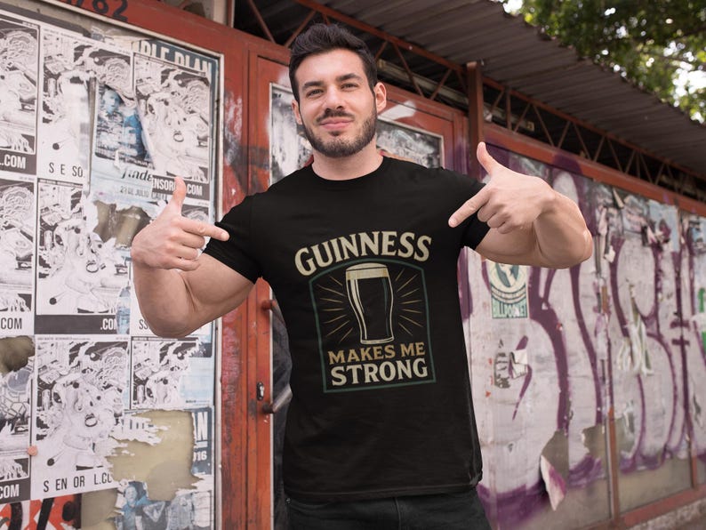 May include: Black t-shirt with the text "GUINNESS MAKES ME STRONG" in a cream-colored font. The design includes an illustration of a pint of Guinness. The person in the image is pointing at the shirt.