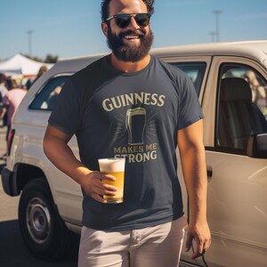May include: Navy blue t-shirt with the Guinness logo and the text "Guinness Makes Me Strong." The shirt is paired with khaki shorts. The person is holding a glass of beer.