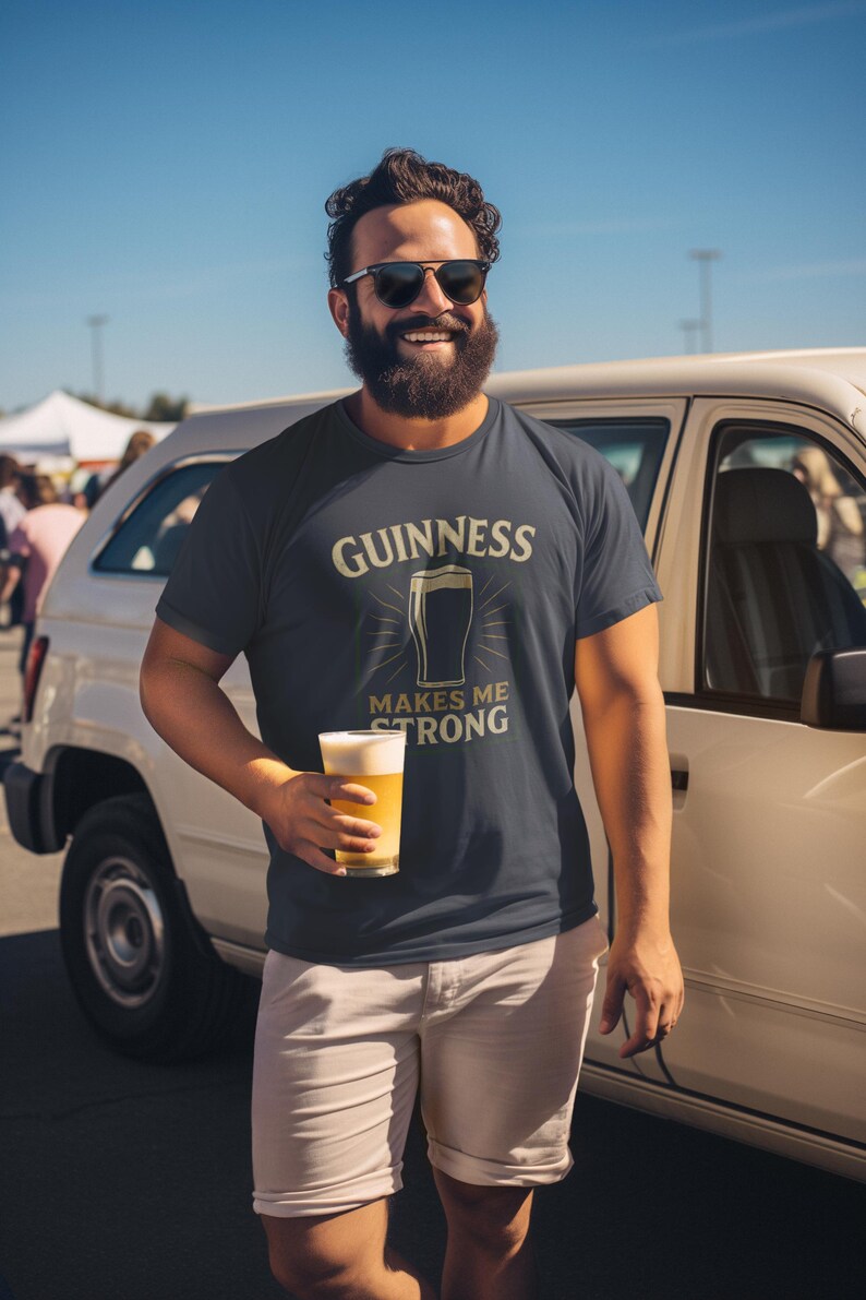 May include: Navy blue t-shirt with the Guinness logo and the text "Guinness Makes Me Strong." The shirt is paired with khaki shorts. The person is holding a glass of beer.