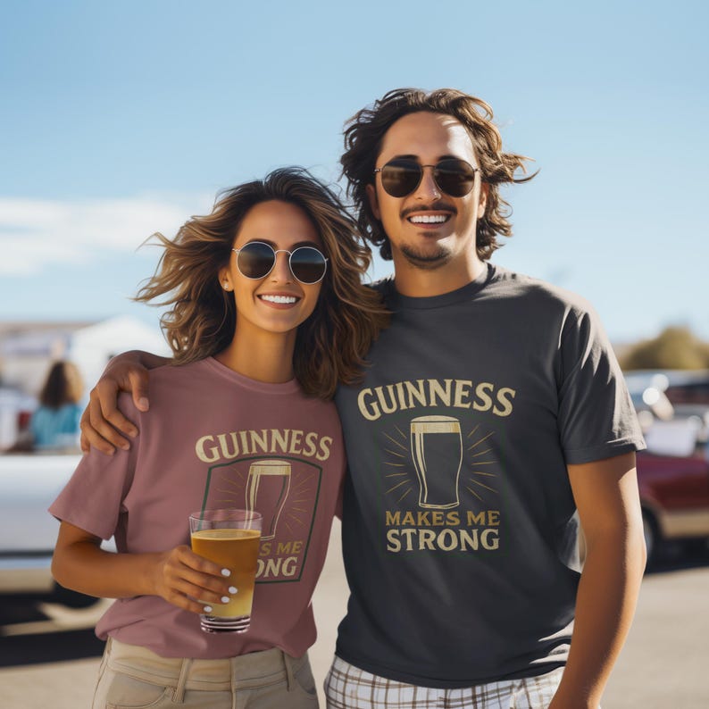 May include: Two people wearing matching t-shirts with the Guinness logo and a pint of beer. The shirts are in different colors, one is a dusty rose color and the other is dark gray. The text on the shirts says "Guinness Makes Me Strong".