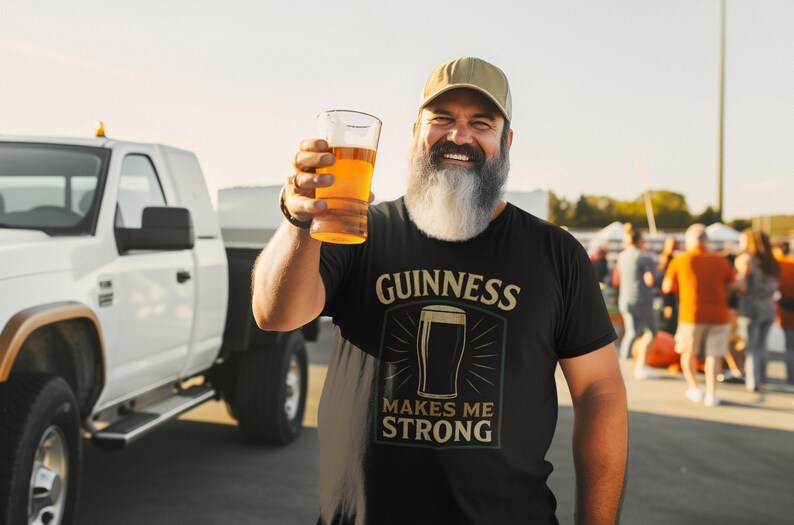 May include: A man wearing a black t-shirt with the Guinness logo and the text "MAKES ME STRONG" holds up a glass of beer. He is wearing a baseball cap and has a long, gray beard. A white truck is in the background.