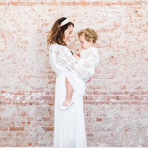 May include: A woman and a young girl wearing white lace dresses. The woman is holding the girl in her arms. They are standing in front of a brick wall.