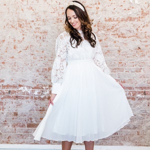 May include: A white midi dress with long sleeves and a lace overlay. The dress has a pleated skirt and a fitted bodice. The dress is paired with white heels.