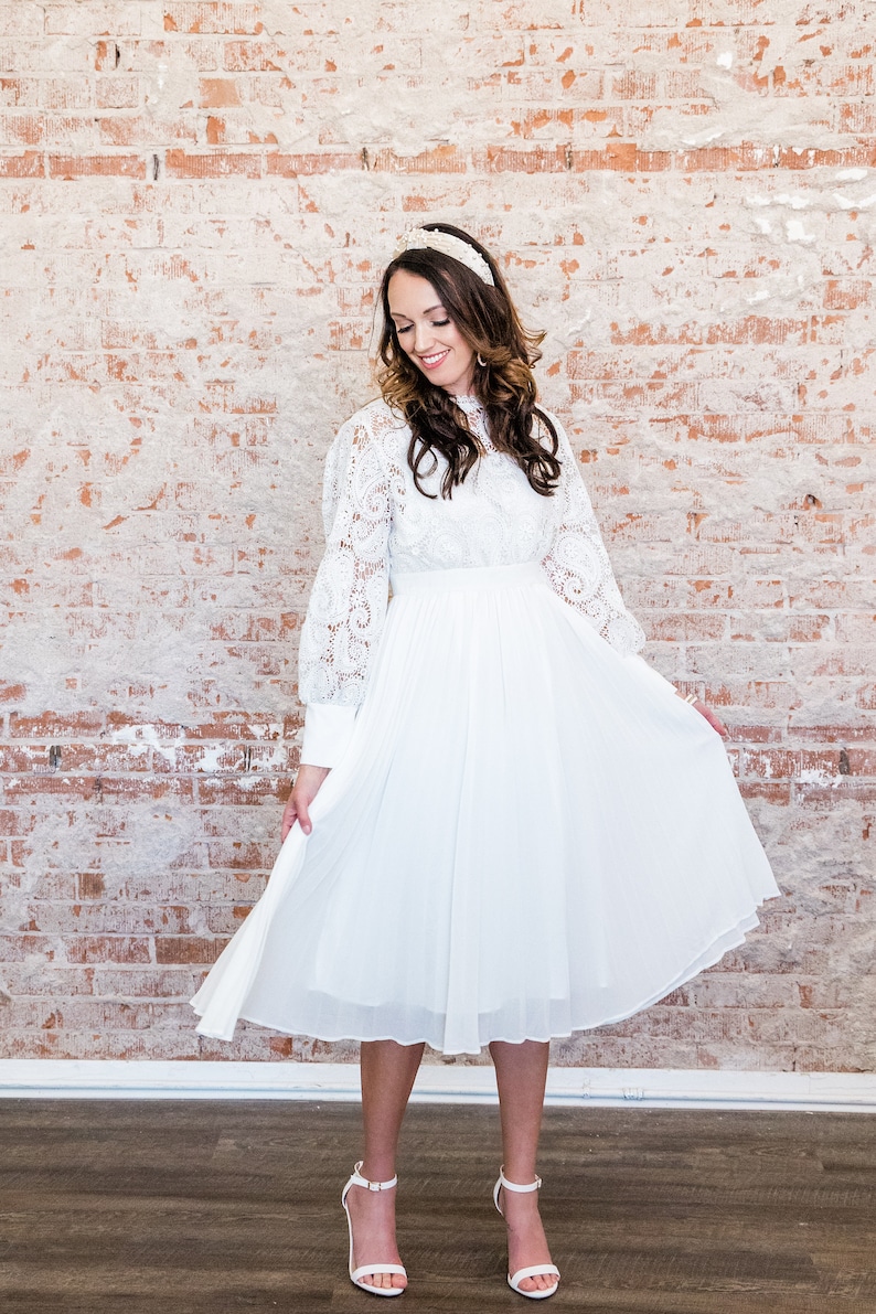 May include: A white midi dress with long sleeves and a lace overlay. The dress has a pleated skirt and a fitted bodice. The dress is paired with white heels.