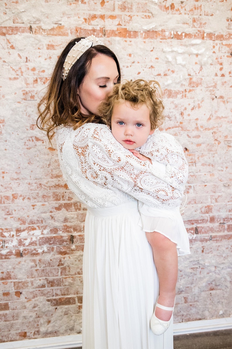 May include: A woman in a white lace dress holds a young child in a white dress. The woman is wearing a pearl headband.