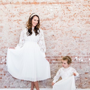 May include: Two people wearing white lace dresses. The person on the left is an adult and the person on the right is a child. The adult is holding the skirt of her dress and smiling at the camera. The child is holding the skirt of her dress and looking at the camera. The background is a brick wall.