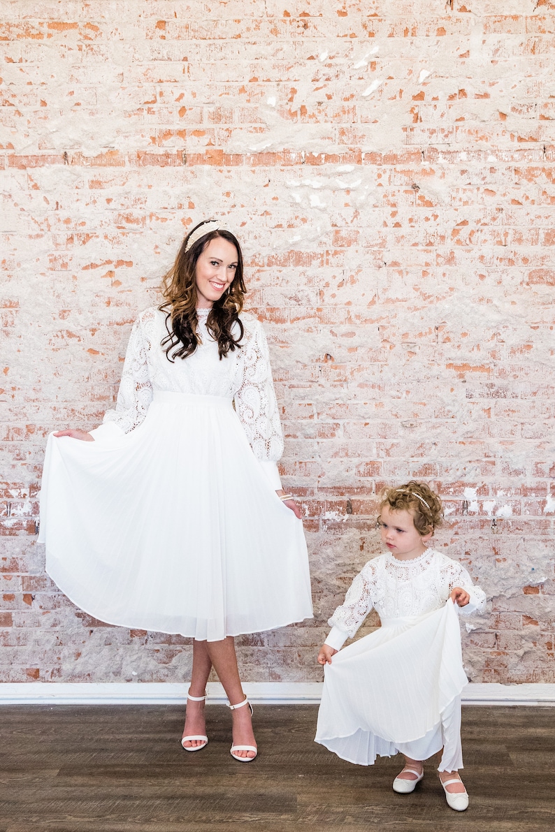 May include: Two people wearing white lace dresses. The person on the left is an adult and the person on the right is a child. The adult is holding the skirt of her dress and smiling at the camera. The child is holding the skirt of her dress and looking at the camera. The background is a brick wall.