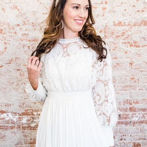 May include: A white lace dress with long sleeves and a pleated skirt. The dress has a high neckline and a fitted bodice. The dress is paired with a pearl headband and pearl bracelets.