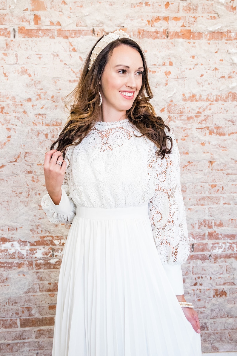 May include: A white lace dress with long sleeves and a pleated skirt. The dress has a high neckline and a fitted bodice. The dress is paired with a pearl headband and pearl bracelets.