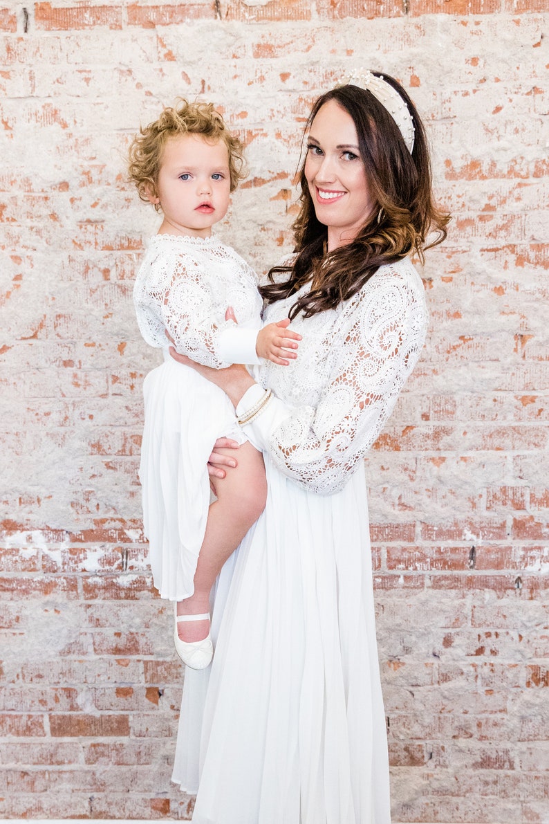 May include: A woman and a child wearing white lace dresses. The woman is holding the child in her arms. The child is wearing white shoes.