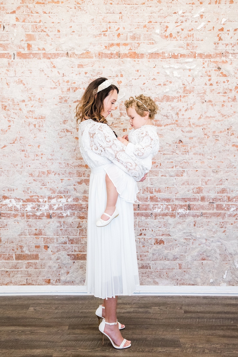 May include: A woman and a young girl wearing white lace dresses. The woman is holding the girl in her arms. They are standing in front of a brick wall.