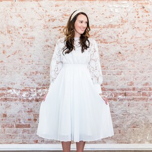May include: A white lace dress with long sleeves and a pleated skirt. The dress is knee-length and has a fitted bodice. The model is wearing white heels.