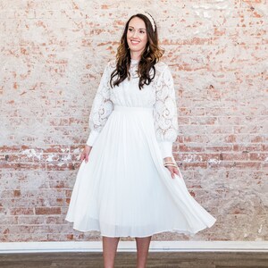 May include: A white lace dress with a pleated skirt and long sleeves. The dress is midi length and has a fitted bodice. The model is wearing white heels.