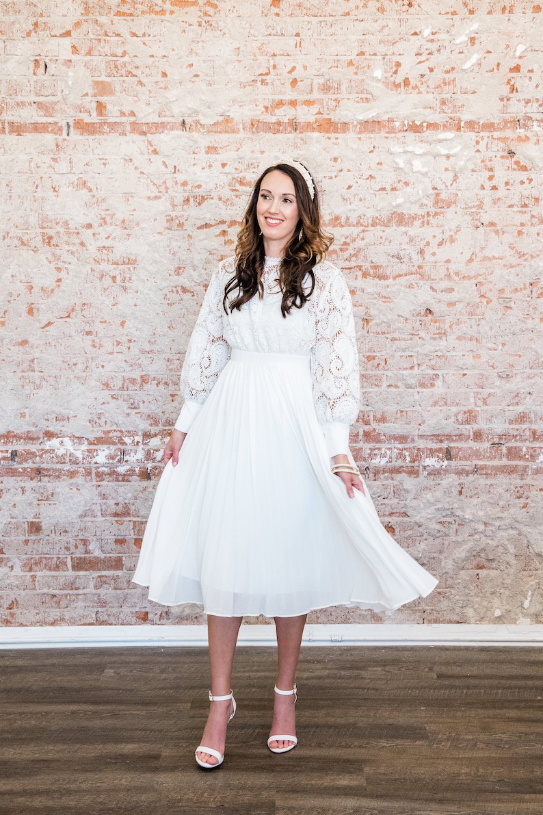 May include: A white lace dress with a pleated skirt and long sleeves. The dress is midi length and has a fitted bodice. The model is wearing white heels.