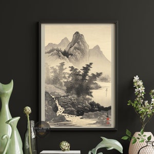Puede incluir: Una impresión en blanco y negro enmarcada de una pintura japonesa de paisaje que representa montañas, árboles y un río. La impresión está colgada en una pared negra con un jarrón de flores blancas y una figura de gato de cerámica verde en primer plano.