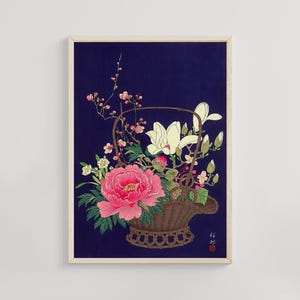 May include: A framed print featuring a floral arrangement in a woven basket against a dark blue background. The arrangement includes a large pink peony, white magnolia blossoms, and delicate pink and white flowers. The basket is brown.