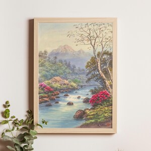 May include: Watercolor painting in a light wood frame. The artwork depicts a serene landscape with a river flowing through a valley, a small house, and a bridge. Lush greenery and vibrant pink flowers frame the water.