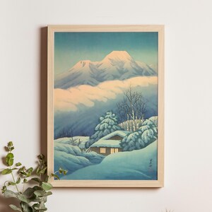 Puede incluir: Impresión enmarcada de un paisaje invernal japonés. La obra muestra montañas nevadas y una pequeña casa. Predominan los colores azul y blanco, con luz cálida de las ventanas. El marco es de madera clara.
