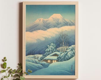 Può includere: Stampa incorniciata di un paesaggio invernale giapponese. L'opera mostra montagne innevate e una piccola casa. I colori dominanti sono blu e bianco, con luce calda dalle finestre. La cornice è in legno chiaro.