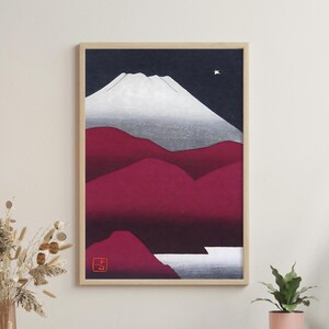 Puede incluir: Impresión artística enmarcada que representa un paisaje de montaña. La obra muestra una montaña nevada sobre un cielo azul oscuro con una estrella. El primer plano muestra colinas de color rojo intenso y una masa de agua blanca. La impresión está en un marco de madera clara.