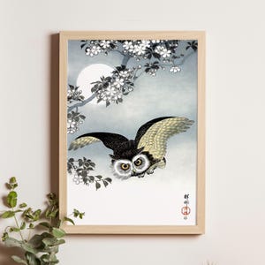May include: A framed print featuring a Japanese-style illustration of an owl in flight. The owl has black and tan feathers, large yellow eyes, and outstretched wings. The background includes a full moon, cherry blossoms, and Japanese text.