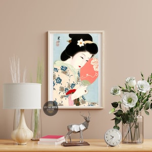 K&ouml;nnte beinhalten: Ein gerahmter Druck einer japanischen Frau in einem traditionellen Kimono, die einen roten F&auml;cher h&auml;lt. Die Frau hat schwarzes Haar und tr&auml;gt eine wei&szlig;e Blume im Haar. Der Hintergrund ist hellblau.