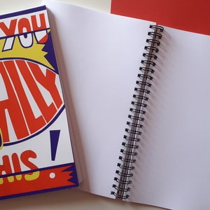 以下が含まれることがあります： 「Can You Totally Do This!」の文字が赤、黄、青で書かれた表紙の、スパイラル綴じノート。ノートは開かれており、白いページが並んでいます。赤い背景が見えます。