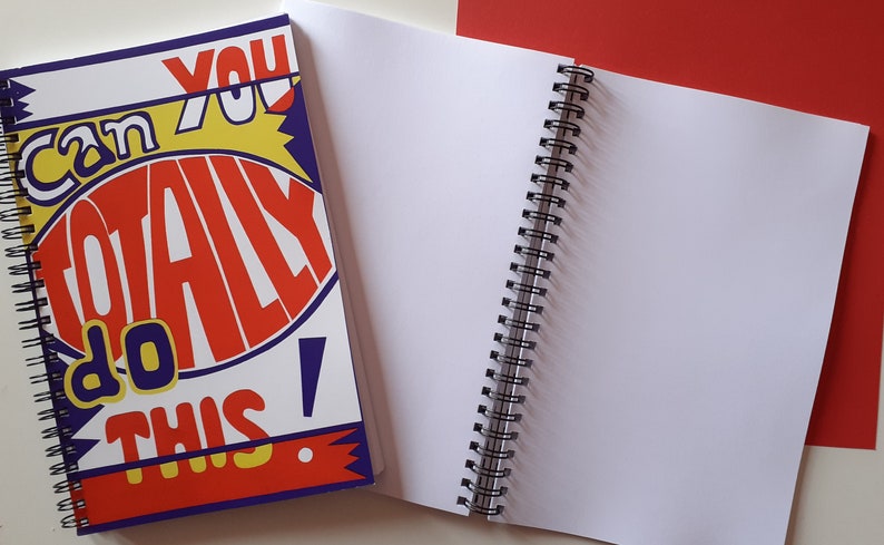 以下が含まれることがあります： 「Can You Totally Do This!」の文字が赤、黄、青で書かれた表紙の、スパイラル綴じノート。ノートは開かれており、白いページが並んでいます。赤い背景が見えます。
