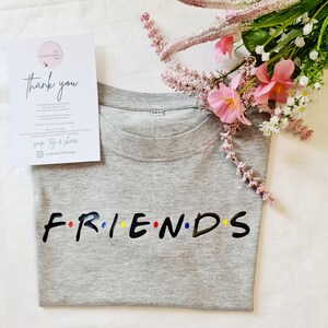 May include: Grey t-shirt with the word "FRIENDS" in black letters with colourful dots between each letter. The shirt is folded and lying on a white surface with pink flowers.