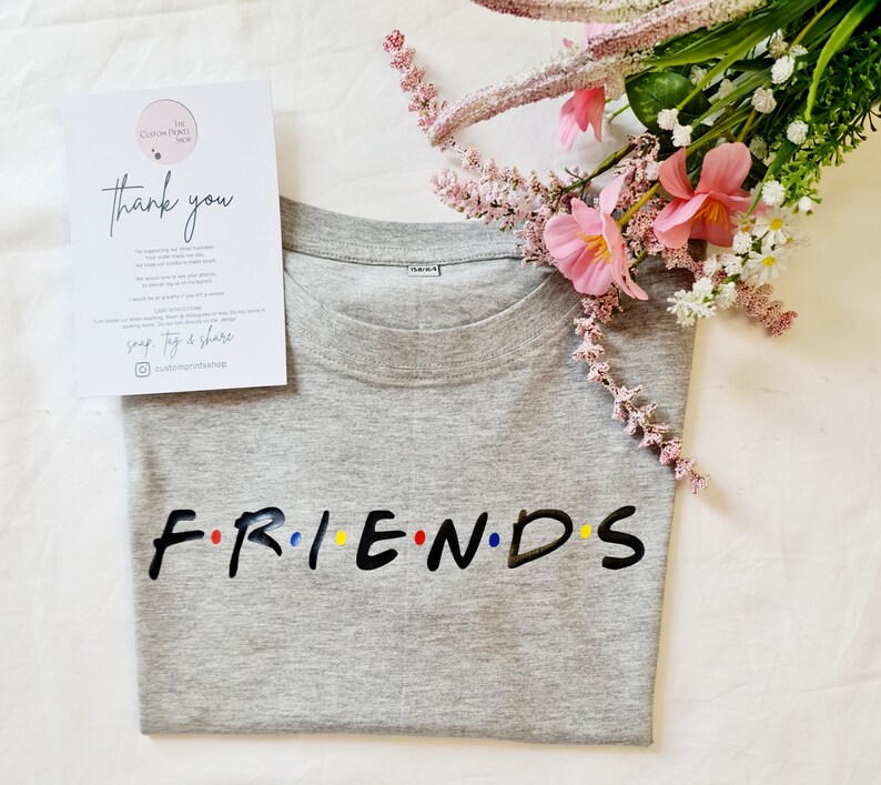 May include: Grey t-shirt with the word "FRIENDS" in black letters with colourful dots between each letter. The shirt is folded and lying on a white surface with pink flowers.