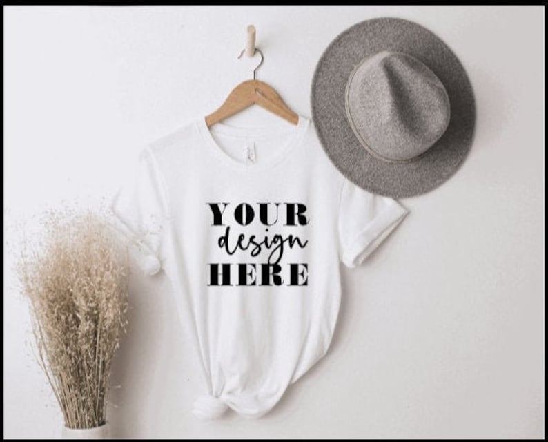 May include: White t-shirt with the text "YOUR design HERE" printed on the front. The shirt is hanging on a wooden hanger against a white wall. A grey hat is lying on the wall to the right of the shirt.