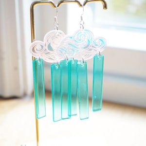 Peut inclure: Boucles d'oreilles en forme de nuages avec des gouttes de pluie rectangulaires pendantes. Les boucles d'oreilles présentent un motif de nuage blanc avec des volutes bleues et des rectangles turquoise transparents.