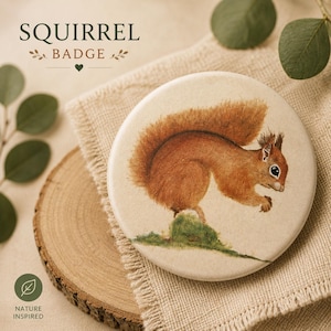 May include: A round badge featuring a detailed illustration of a brown squirrel standing on a patch of green grass. The badge has a cream-coloured background. The text SQUIRREL BADGE is printed in dark green letters above the illustration.