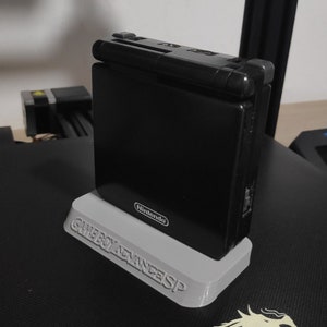 Può includere: Una console Nintendo Game Boy Advance SP nera su un supporto stampato in 3D. Il supporto è etichettato "Game Boy Advance SP" e ha il logo Nintendo.