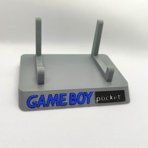 Puede incluir: Un soporte gris para Game Boy Pocket con letras azules. El soporte tiene dos soportes angulados y una base rectangular. El texto "GAME BOY pocket" es visible en la parte delantera.