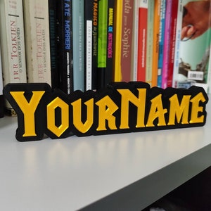 Pode incluir: Uma placa de nome preta e dourada com o texto "YOURNAME" em uma fonte em negrito e estilizada.