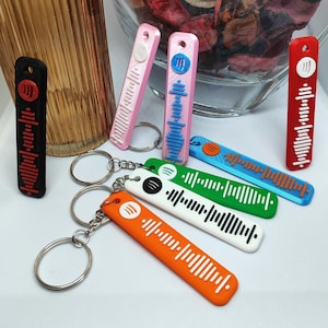 May include: A selection of brightly coloured Spotify code keyrings. The keyrings are in shades of black, pink, orange, white, green, blue, and red. Each has a unique Spotify code design and a silver keyring.