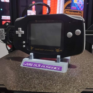 May include: A black Game Boy Advance SP console with a gold Pokemon logo and the text "GAME BOY ADVANCE" on the bottom. The console is sitting on a white 3D printed stand with the text "GAME BOY ADVANCE" in purple.